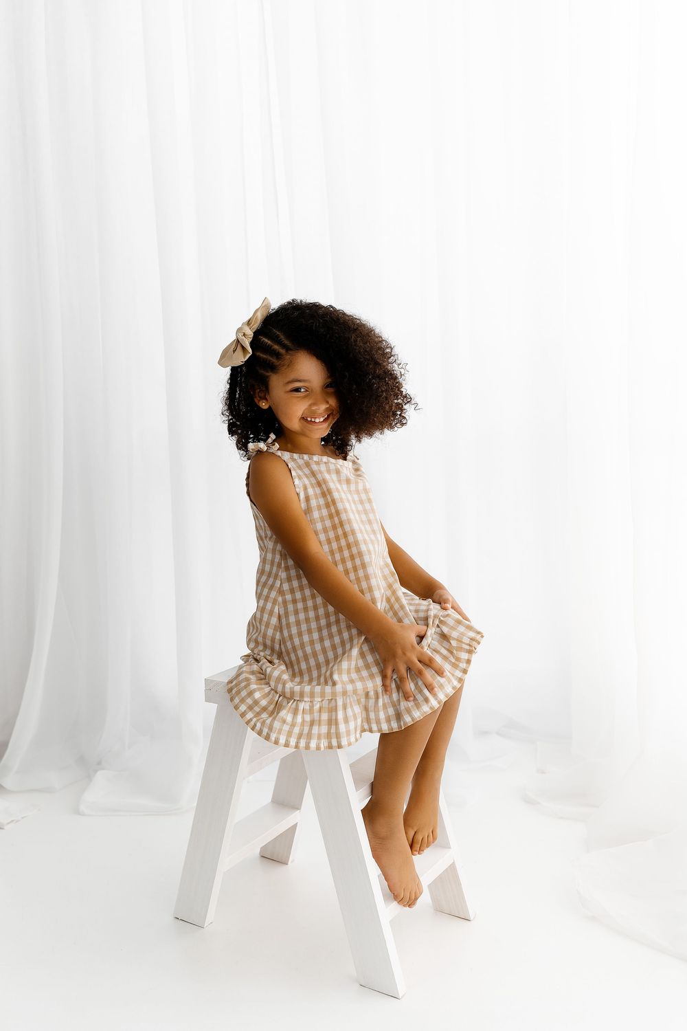 Young child in brown gingham dress with natural curly hair sits on white stool against white backdrop.