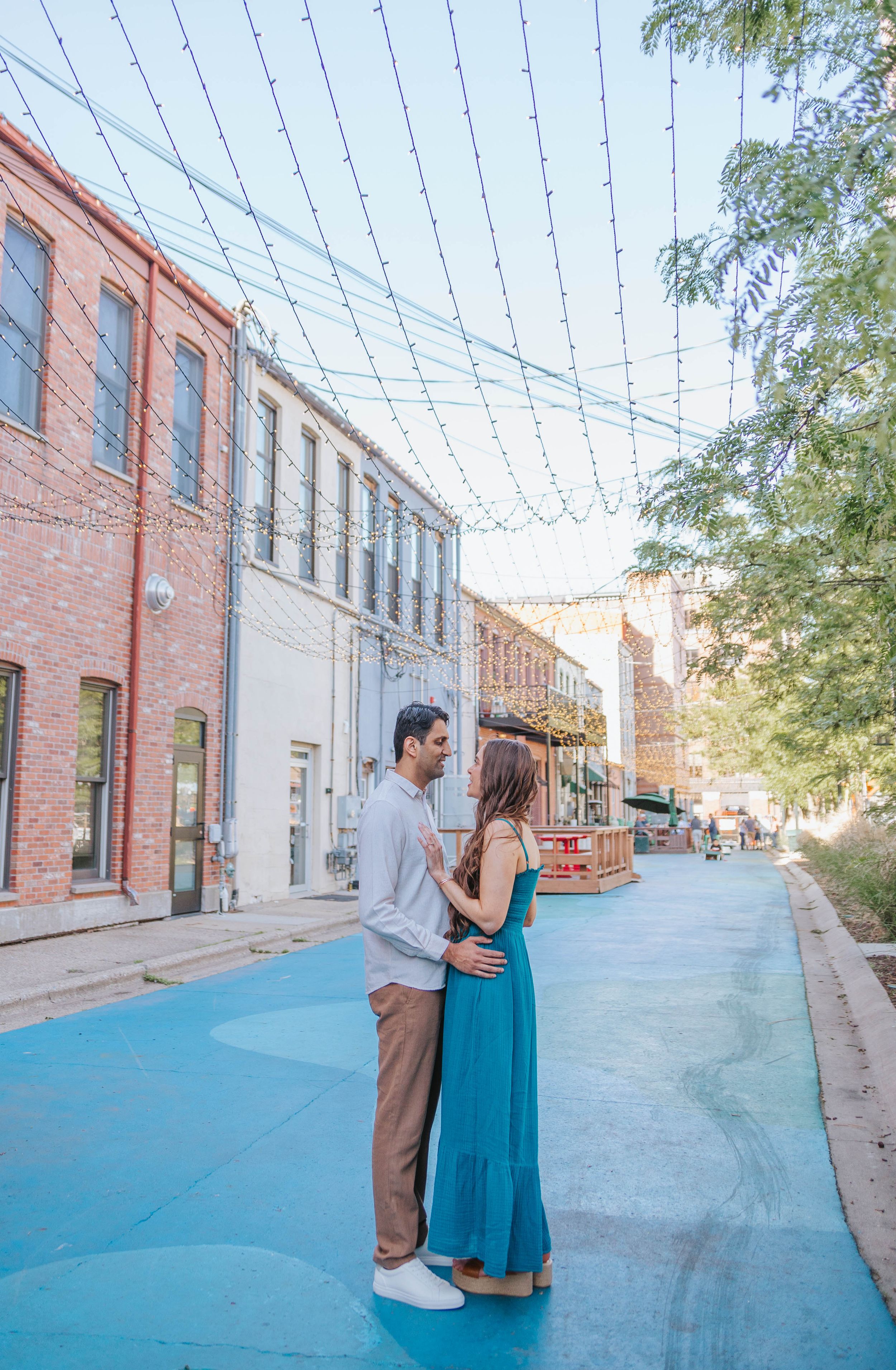 A couple shares an intimate moment on a blue-painted street with string lights and brick buildings overhead.