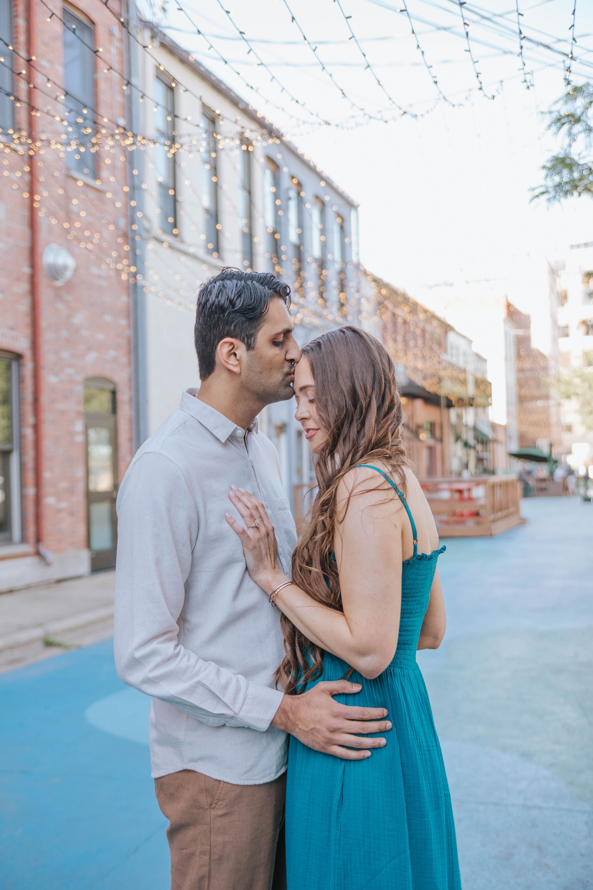 A romantic couple embraces under string lights in front of historic brick buildings on a blue-painted street.