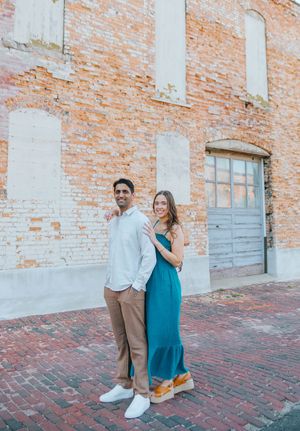 A couple poses together against a rustic brick wall wearing coordinated white and turquoise outfits.