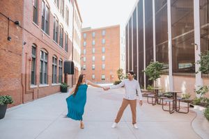 A couple in white and turquoise outfits dances playfully in an urban alleyway.