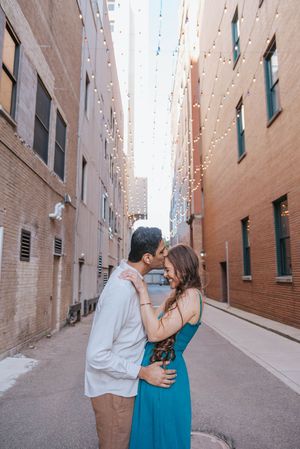 A romantic couple shares an intimate moment in a brick-lined alley wearing blue and beige outfits.