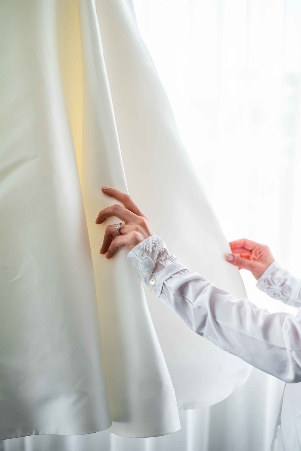Delicate lace-adorned sleeve reaches to adjust a flowing white curtain in soft natural light.