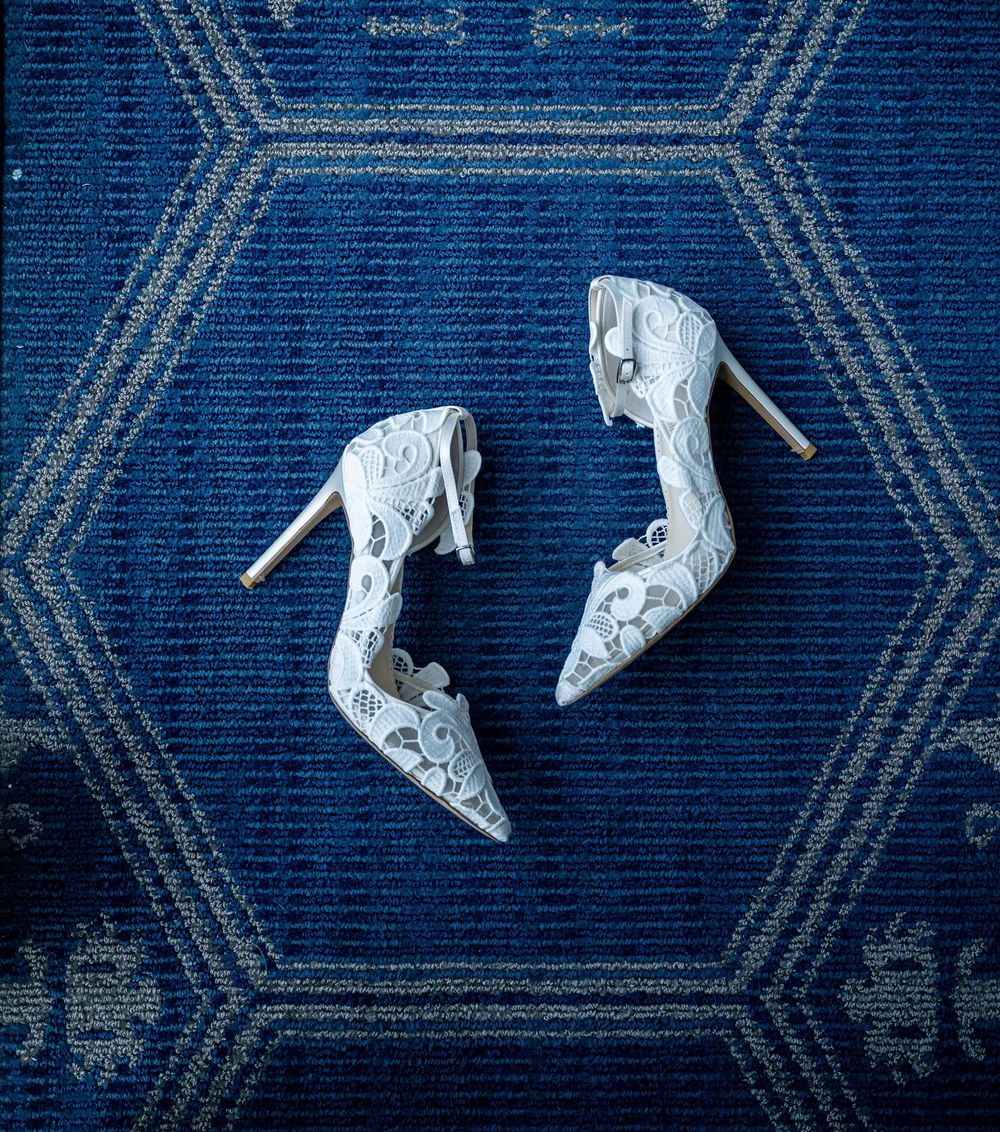 Elegant white lace bridal heels displayed on a blue patterned carpet with geometric designs.