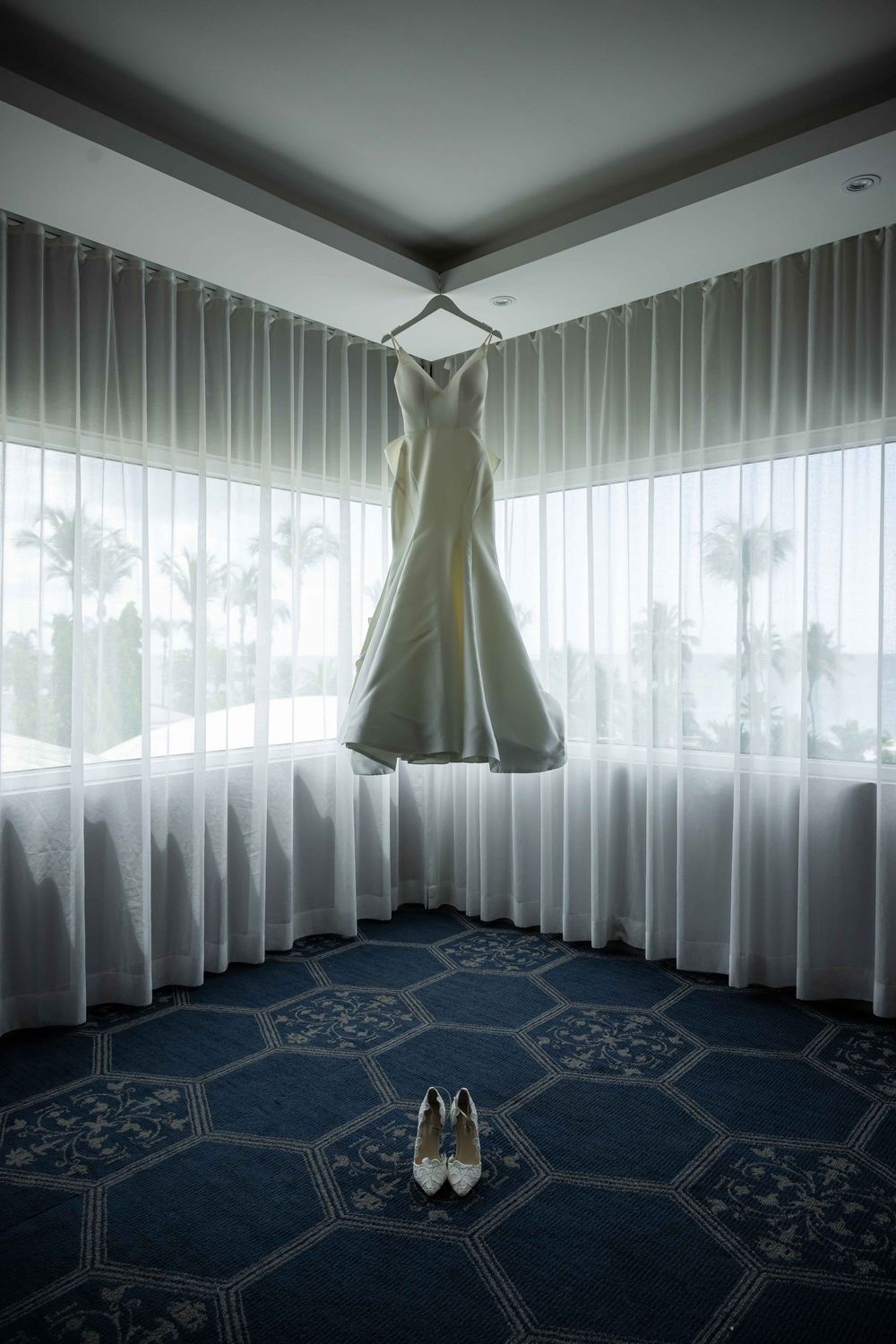 A white wedding dress hangs elegantly in a corner room with sheer curtains and a geometric patterned carpet, with bridal shoes below.