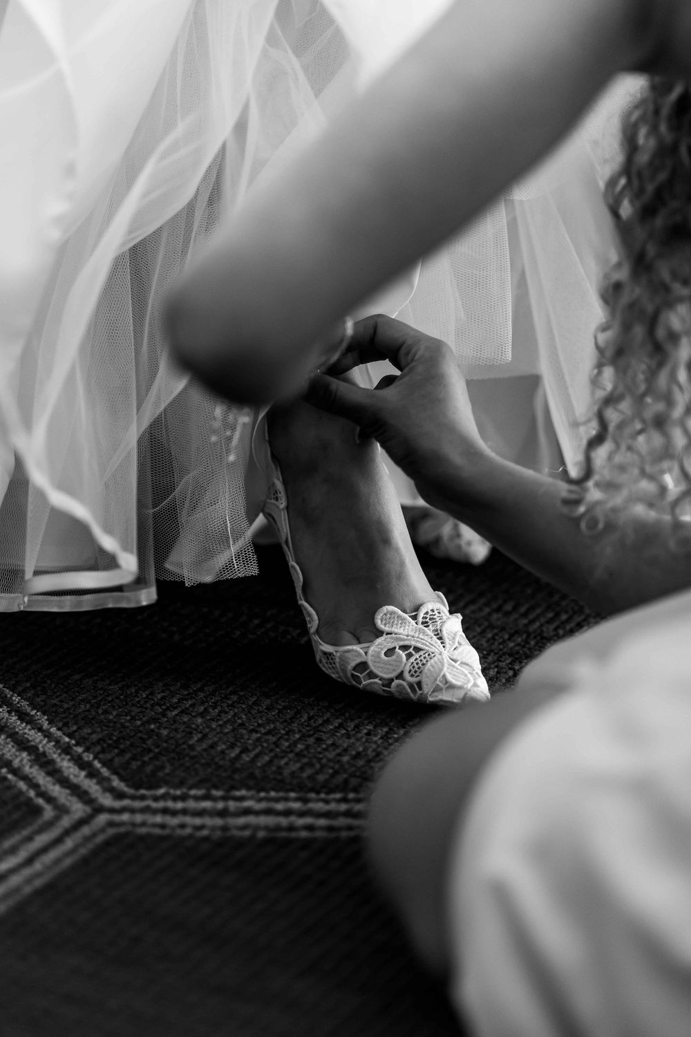 Black and white image of elegant lace bridal shoes being put on during wedding preparations.