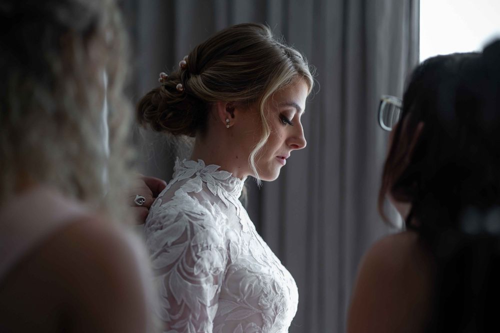A bride in a delicate white lace dress stands in soft window light during wedding preparations.