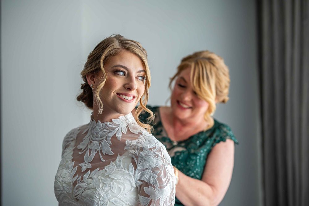 A bride wearing an elegant white lace dress smiles while getting ready with assistance in the background.
