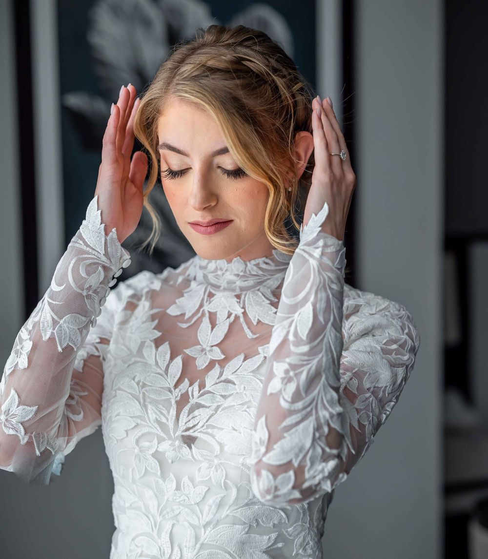 Elegant bride in white lace wedding gown with sheer long sleeves and delicate floral patterns adjusts her hair.