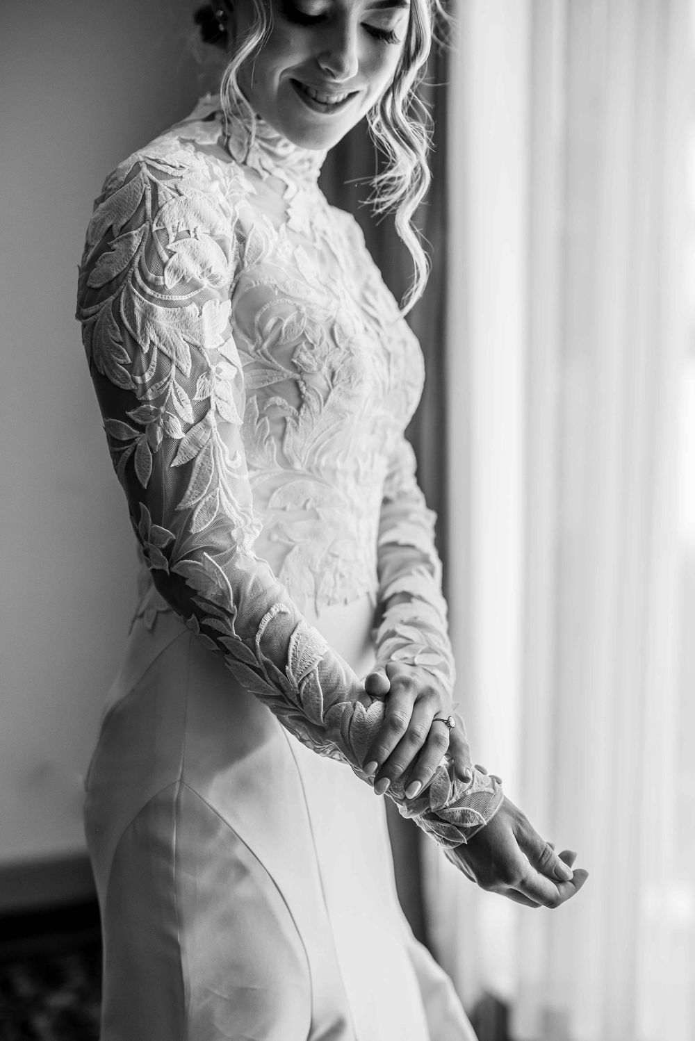 Black and white photo of bride in long sleeve lace wedding dress with delicate details and fitted silhouette by window.