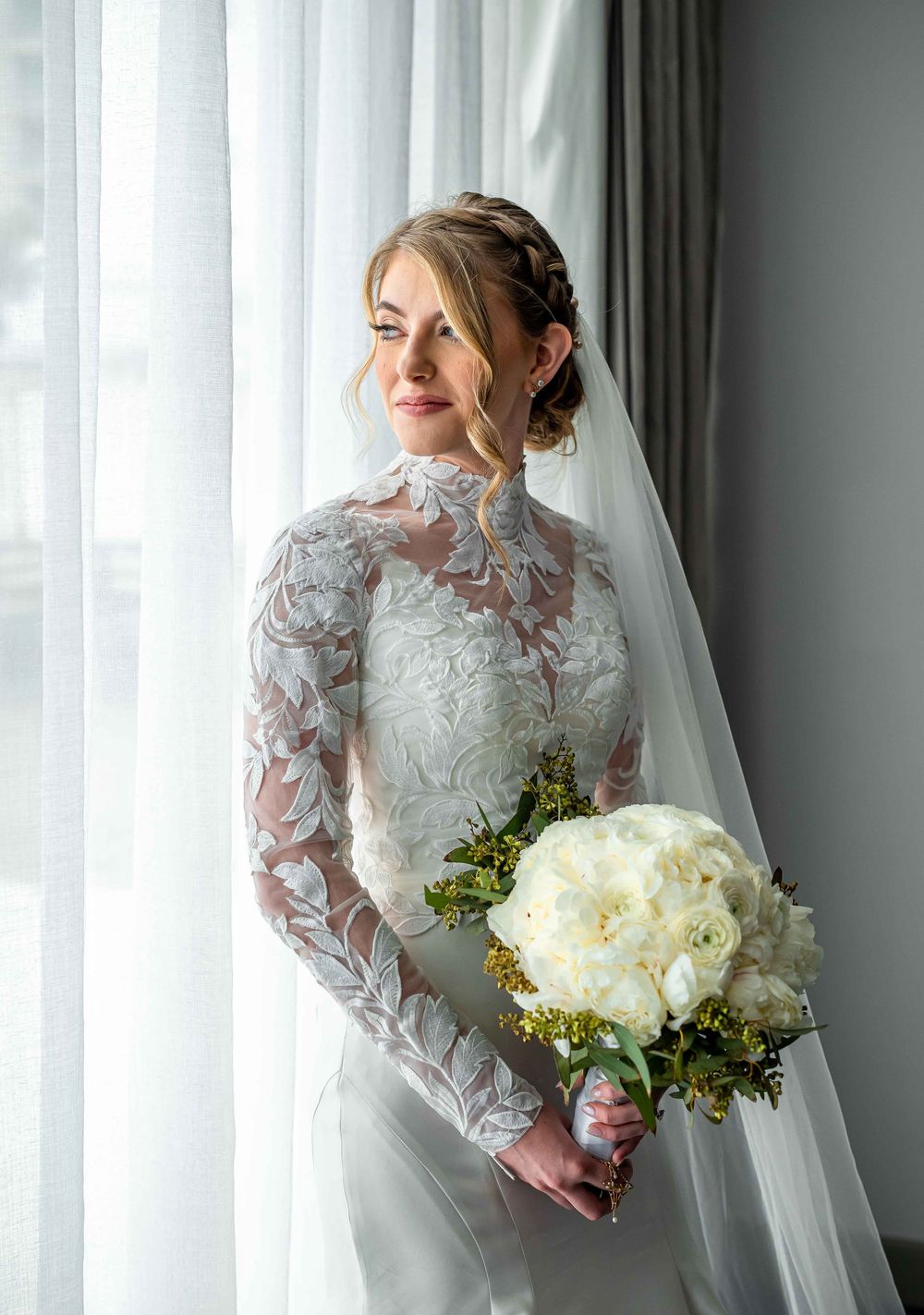 Bride in elegant lace long sleeve wedding dress stands by window holding white peony bouquet on her wedding day.