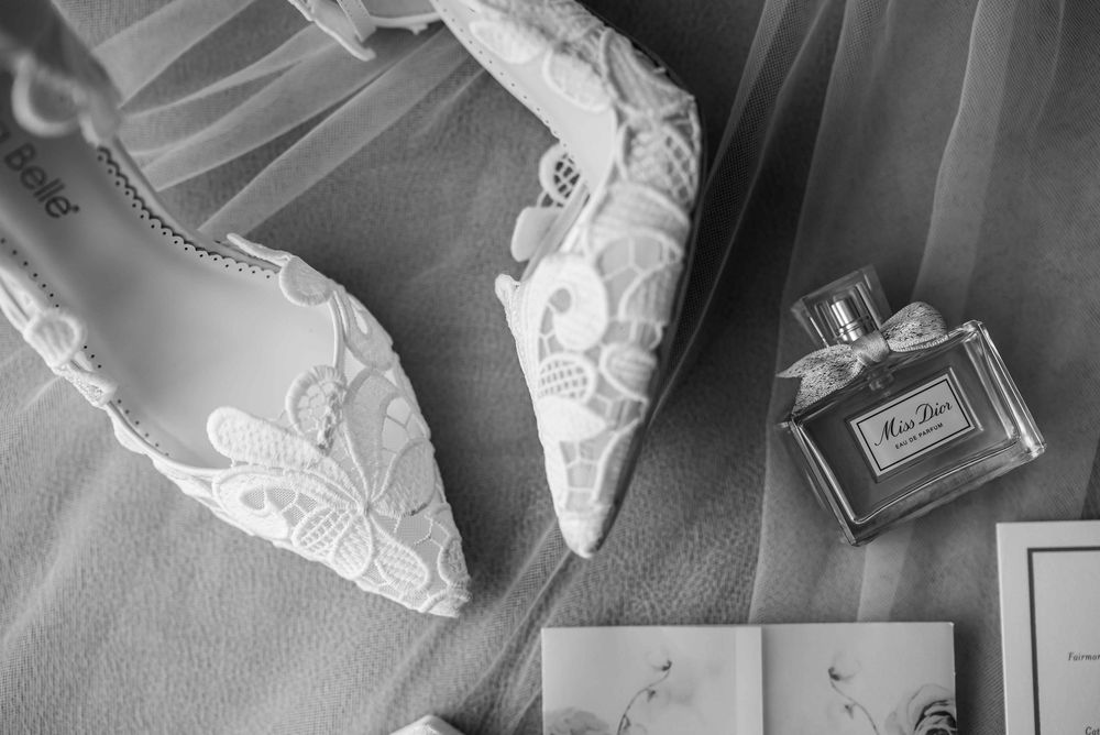 Elegant white lace wedding shoes displayed next to a glass perfume bottle in black and white photography.
