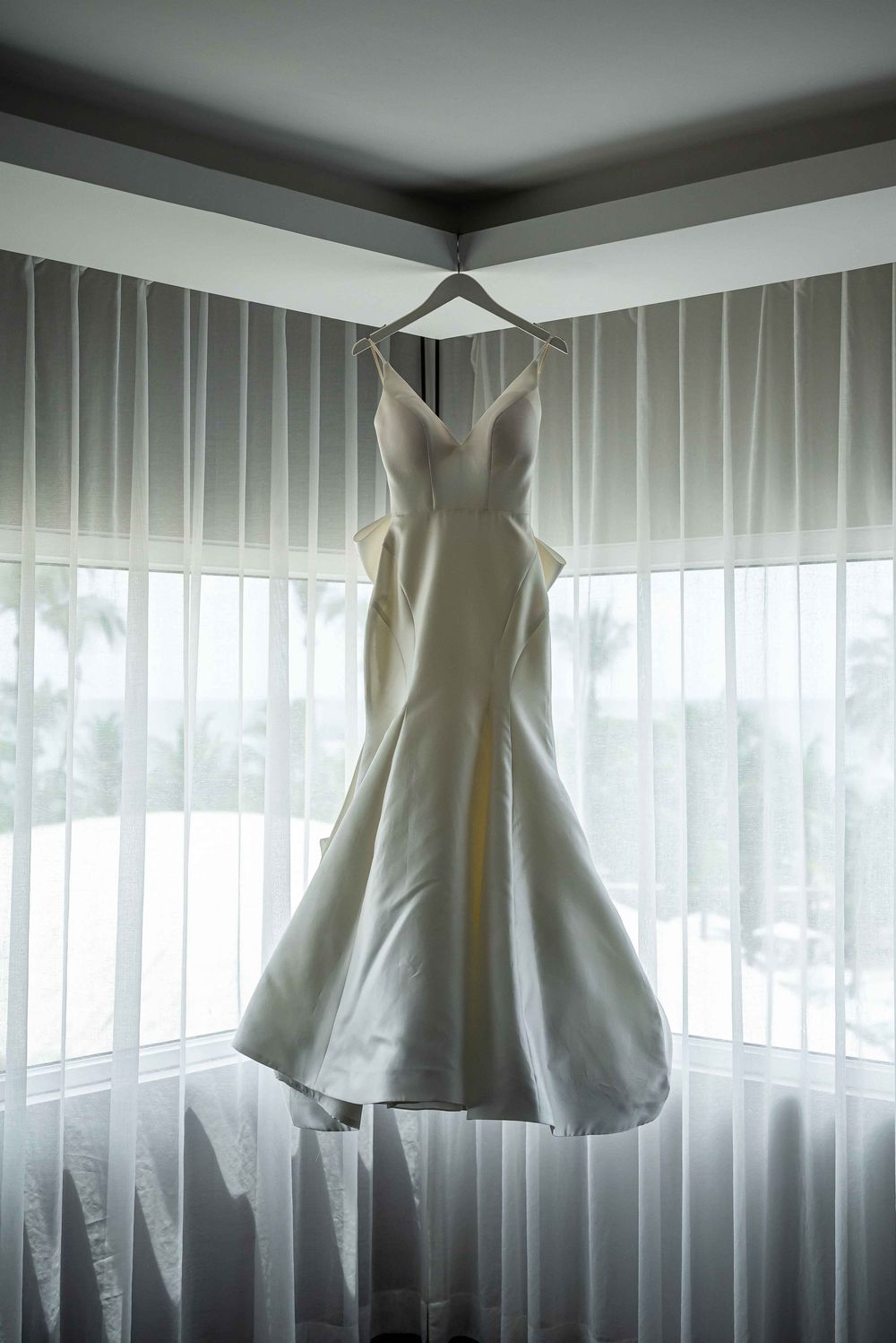 A white wedding dress hangs elegantly from a ceiling with sheer curtains in a modern room with natural lighting.