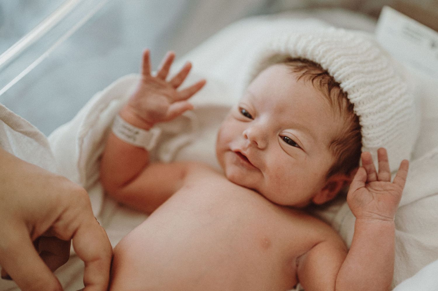 Sleeping newborn baby in soft white bedding with hands up near face, photographed in gentle natural light.