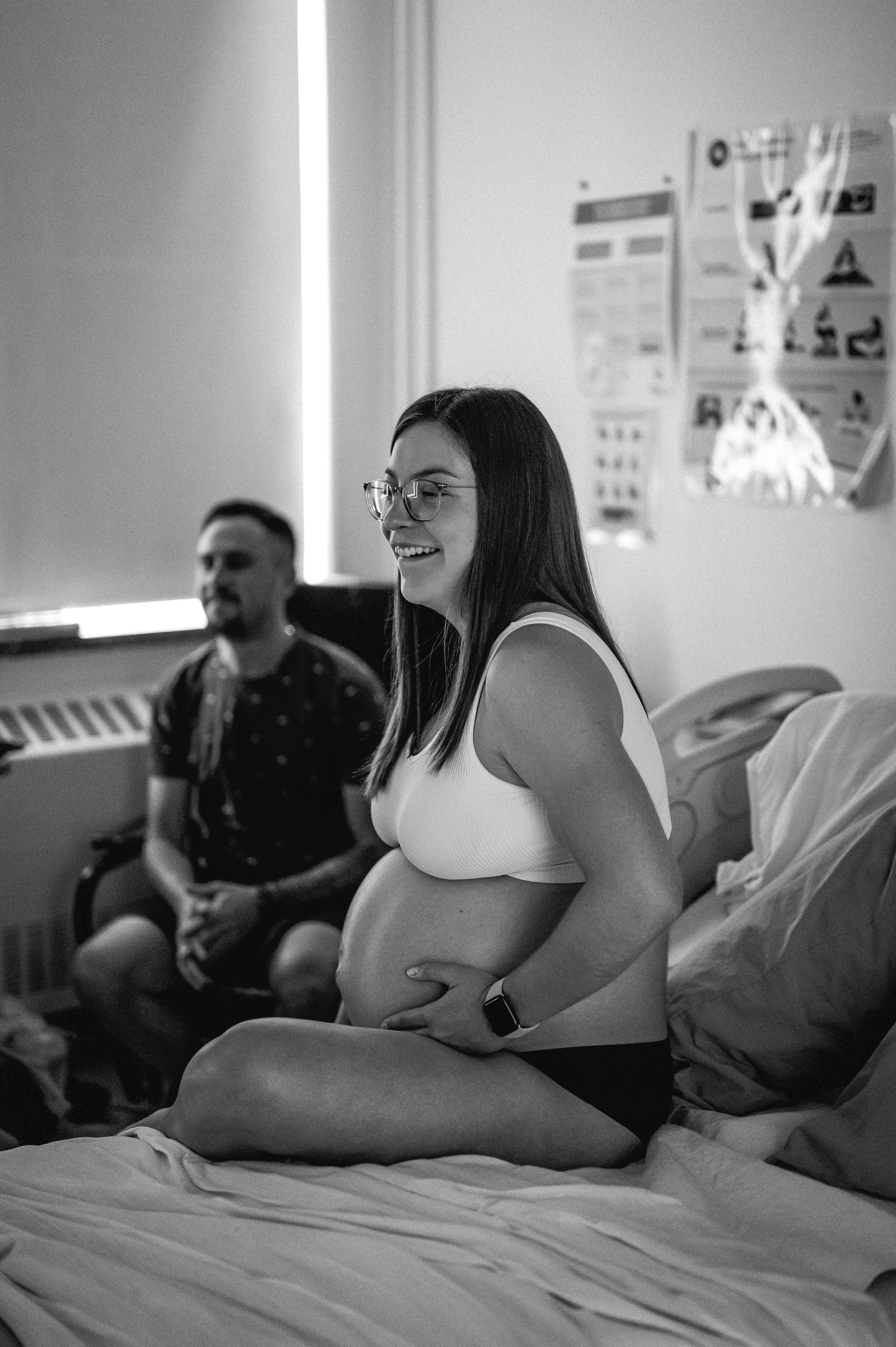 Black and white photo of a pregnant woman sitting cross-legged on a bed while someone sits nearby in the background.