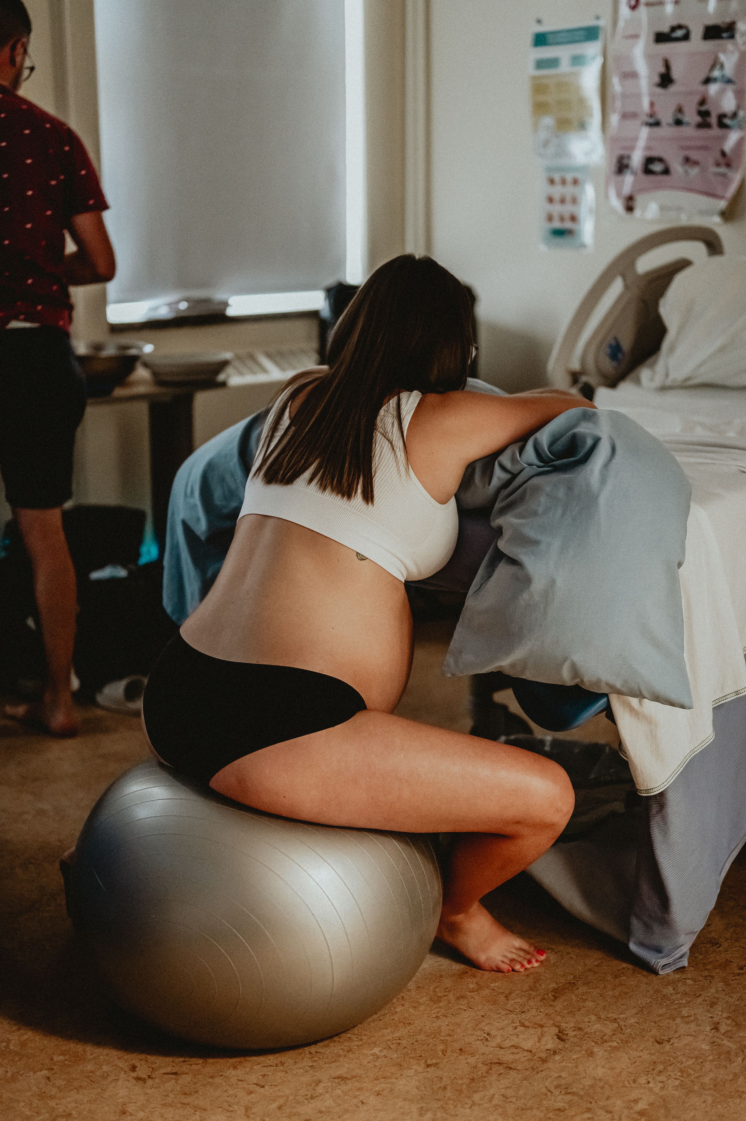 A pregnant woman in exercise wear sits on a gray stability ball during labor or birthing preparation in a medical setting.