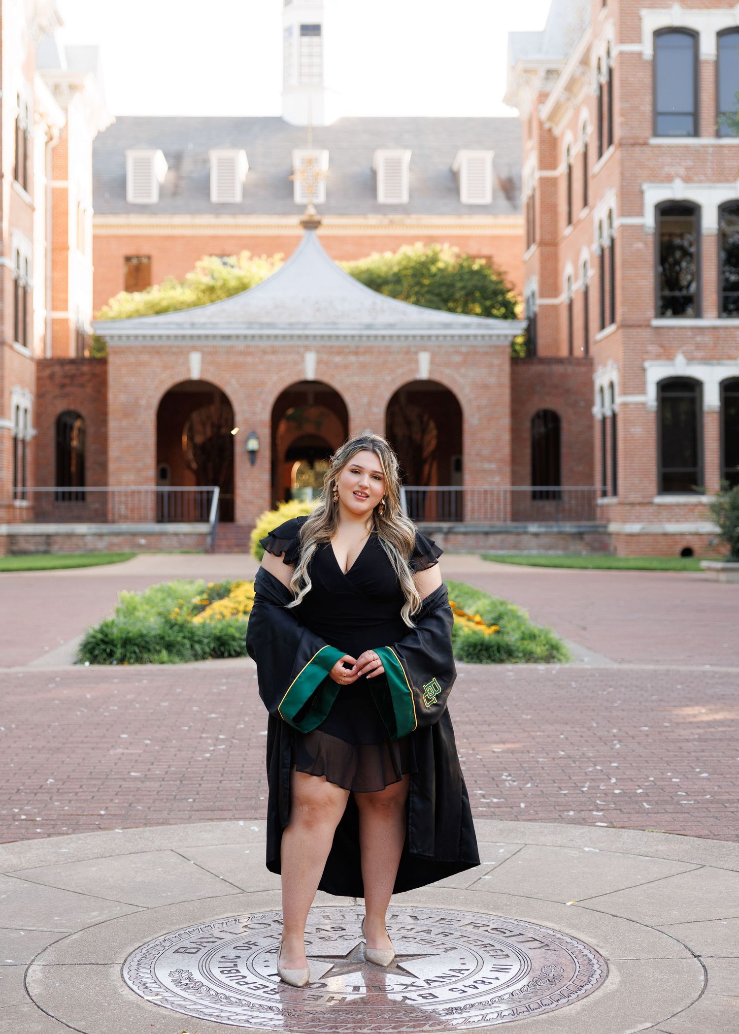 A graduating student in green cap and gown poses for a series of celebratory photos outside on a brick path at Baylor University.