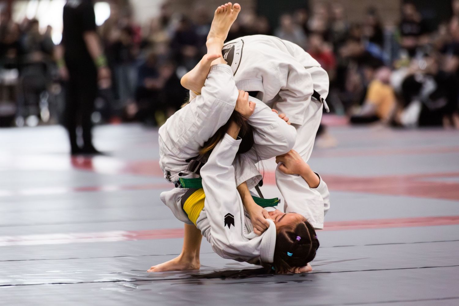 Two Brazilian jiu-jitsu competitors grapple on the mat during a tournament match with spectators in background.