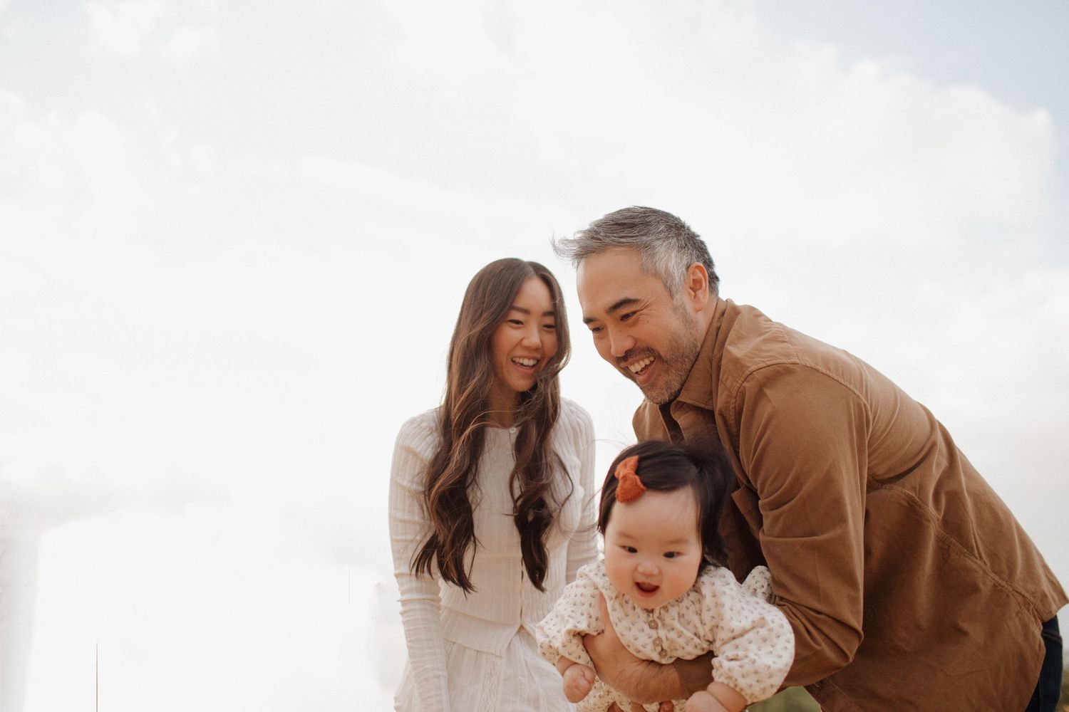 A family shares a joyful moment outdoors together with their baby against a bright sky background.
