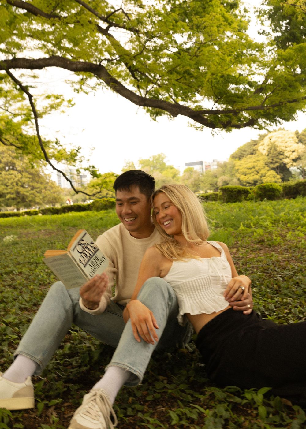 Two people relaxing under a tree reading a book together in a lush green park setting during autumn.
