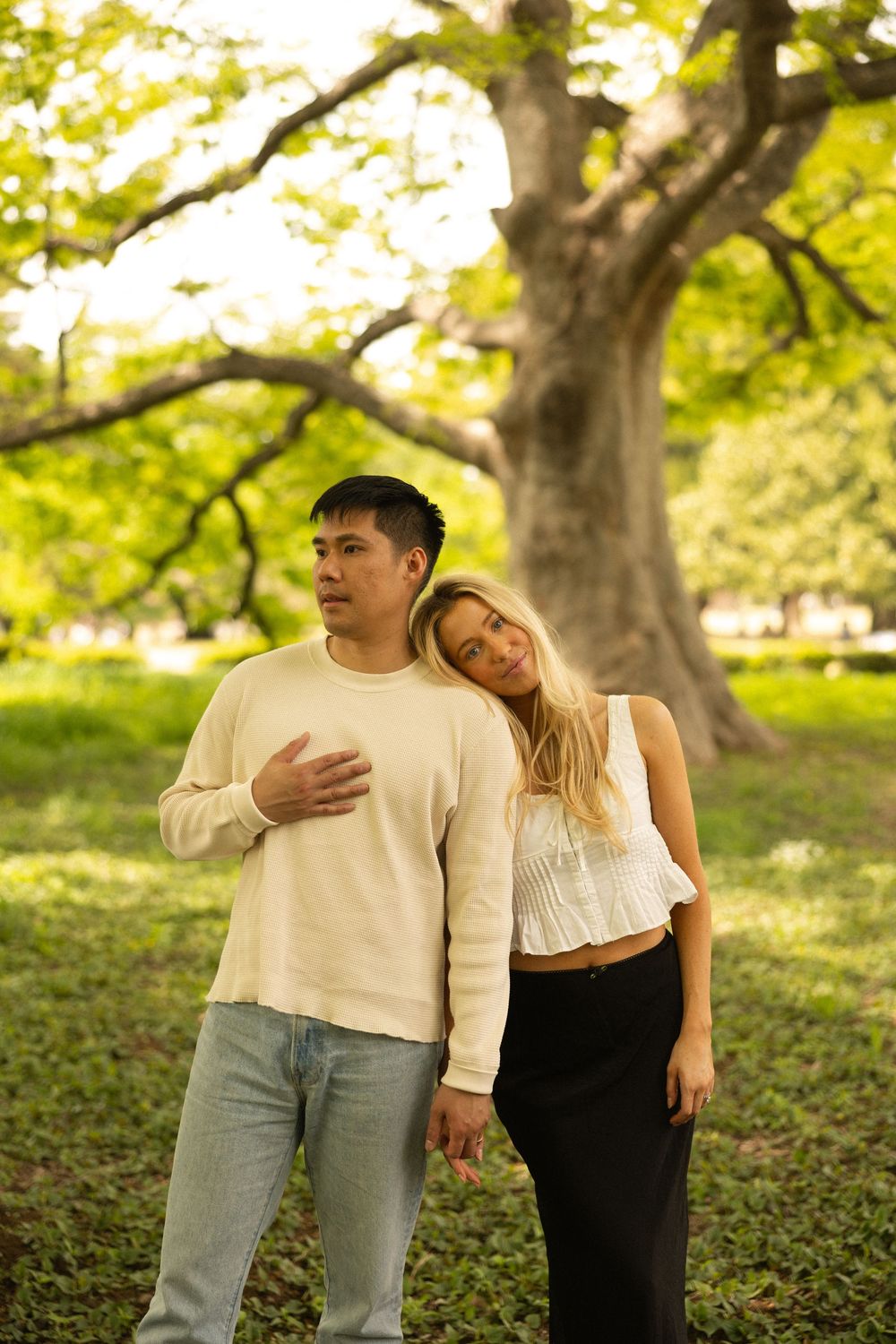 A couple in coordinating cream outfits poses against a sunlit tree in a park during golden hour.