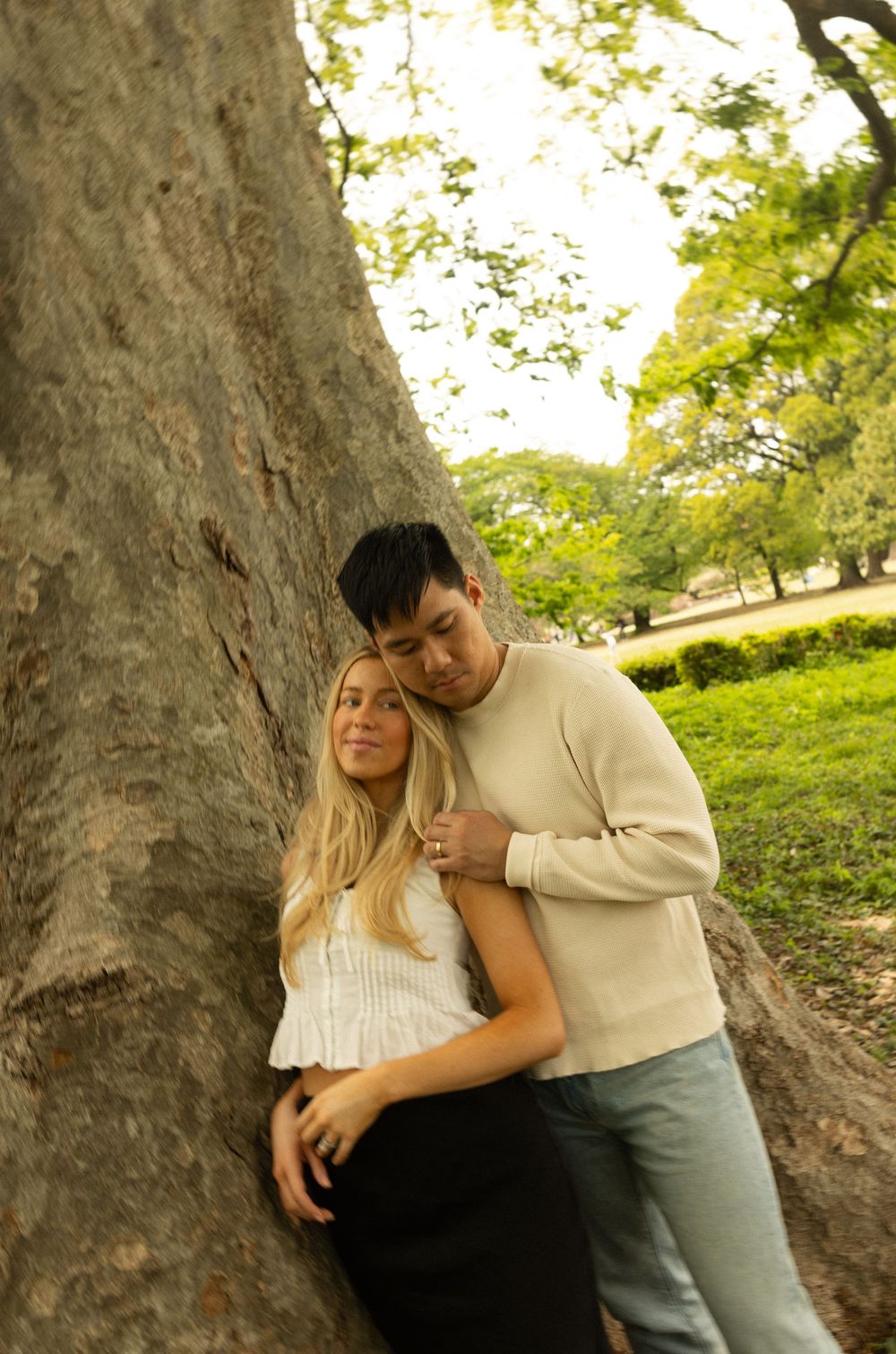 A romantic couple embraces against a large tree trunk in a sunny park setting.