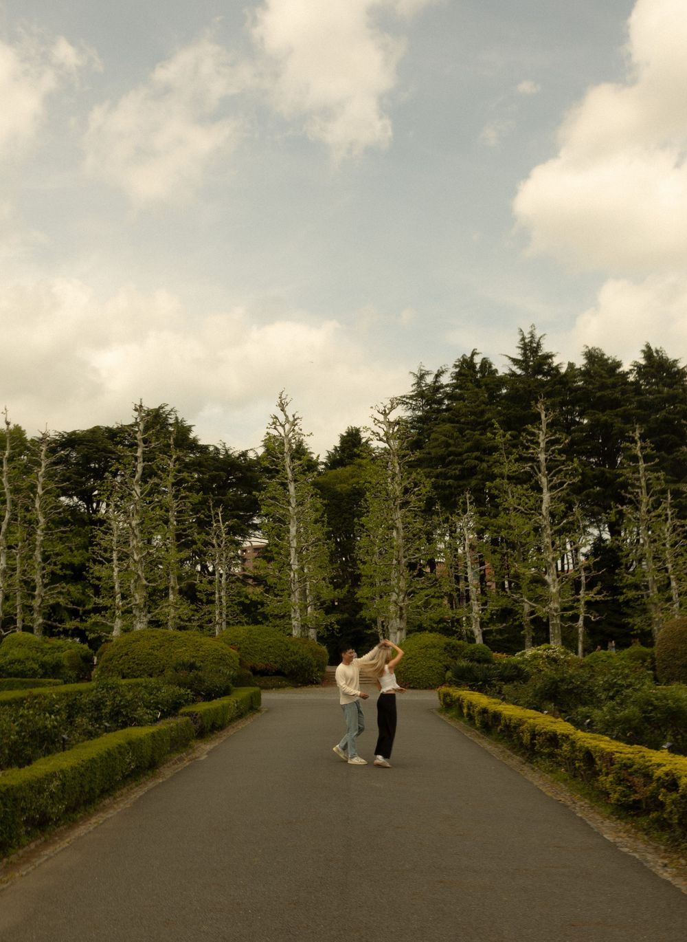 Two people walk along a tree-lined path under cloudy skies in a peaceful garden setting.