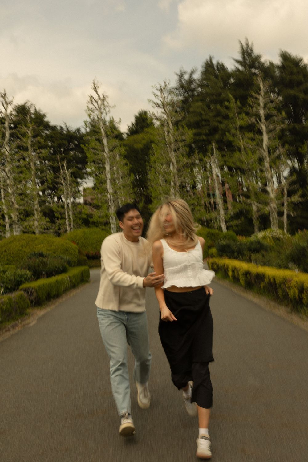 A joyful couple running and laughing together on a tree-lined path under an overcast sky.