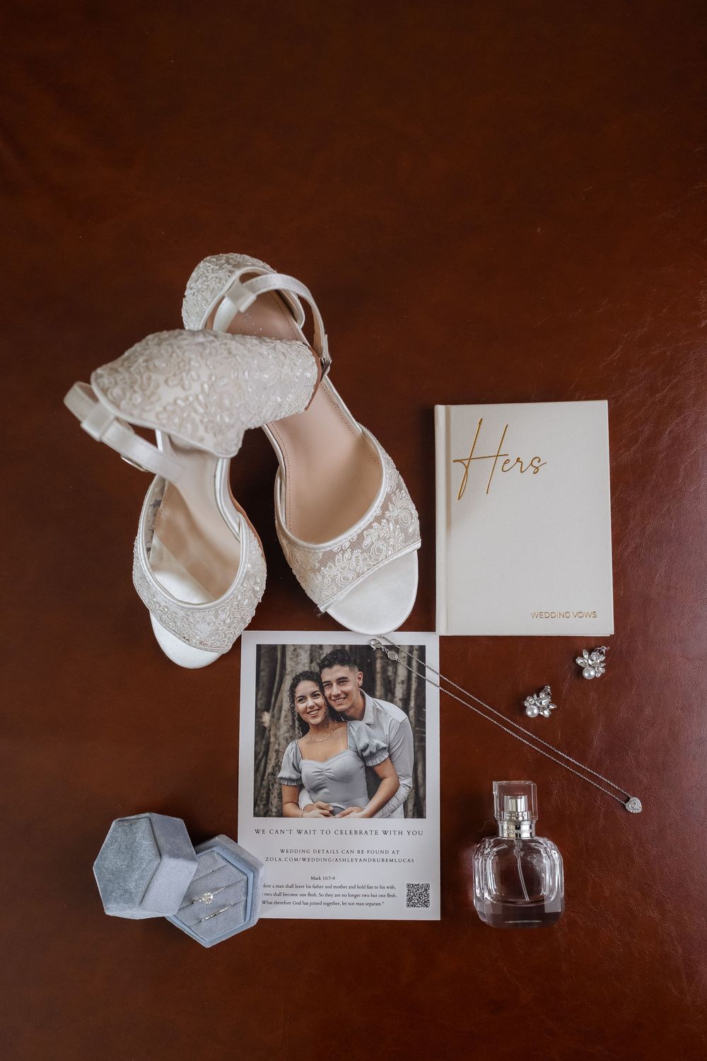 Wedding details flatlay with white lace shoes, perfume bottle, jewelry and invitation card on dark wood surface.