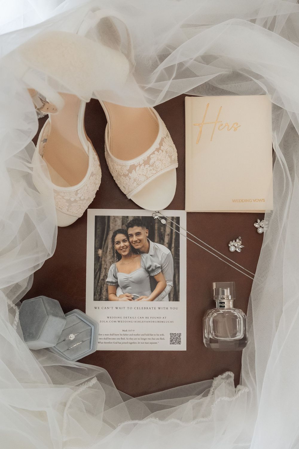 Wedding details flatlay with lace shoes, tulle veil, perfume bottle and save the date card on brown surface.