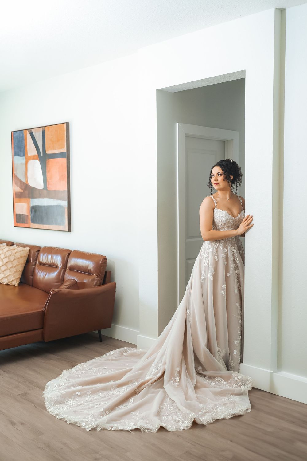 Bride poses in elegant lace wedding gown with flowing train beside brown leather couch.