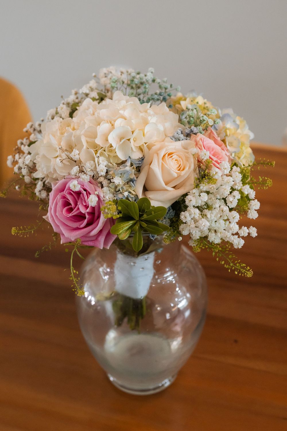 A delicate floral arrangement with pink and cream roses, baby's breath, and greenery in a clear glass vase on a wooden surface.