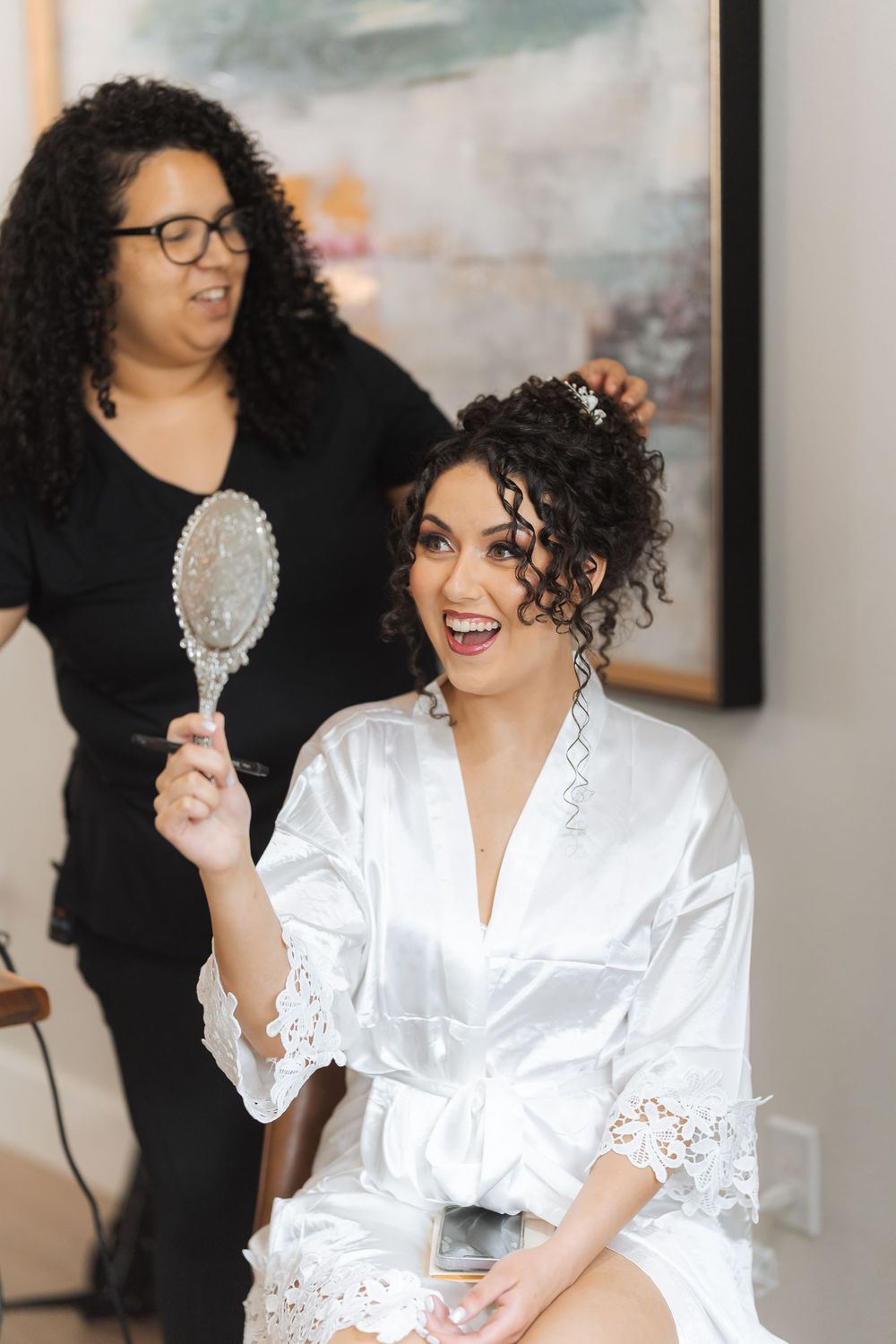 A bride in white satin robe holds a silver mirror during wedding preparations.
