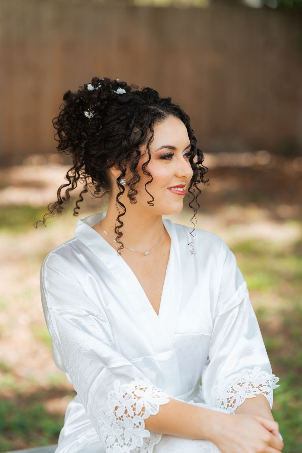 A person in a white robe with lace trim smiles while getting ready, wearing curly hair adorned with small flowers.