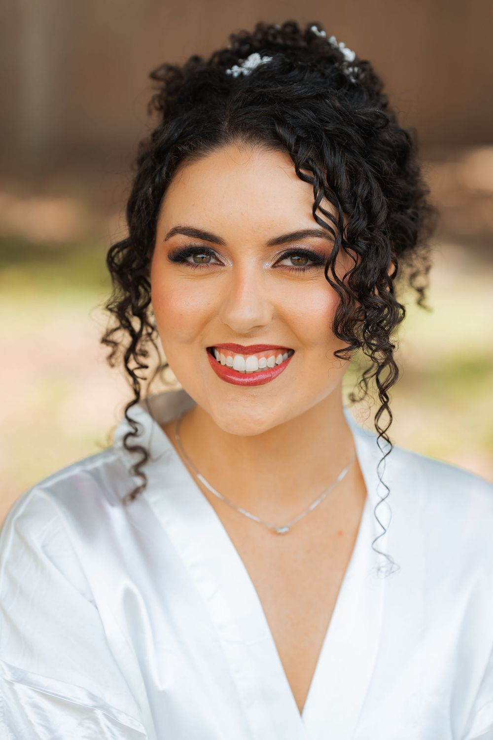 A woman in a white satin robe smiles with curly dark hair styled up and sparkling jewelry.