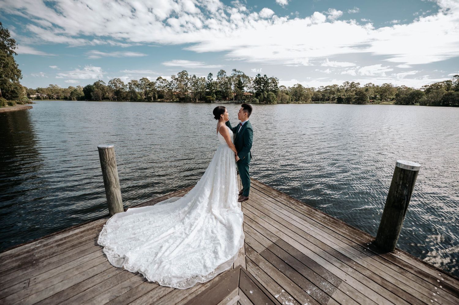 A romantic wedding moment captured on a wooden dock overlooking a serene lake with clouds reflecting on the water.