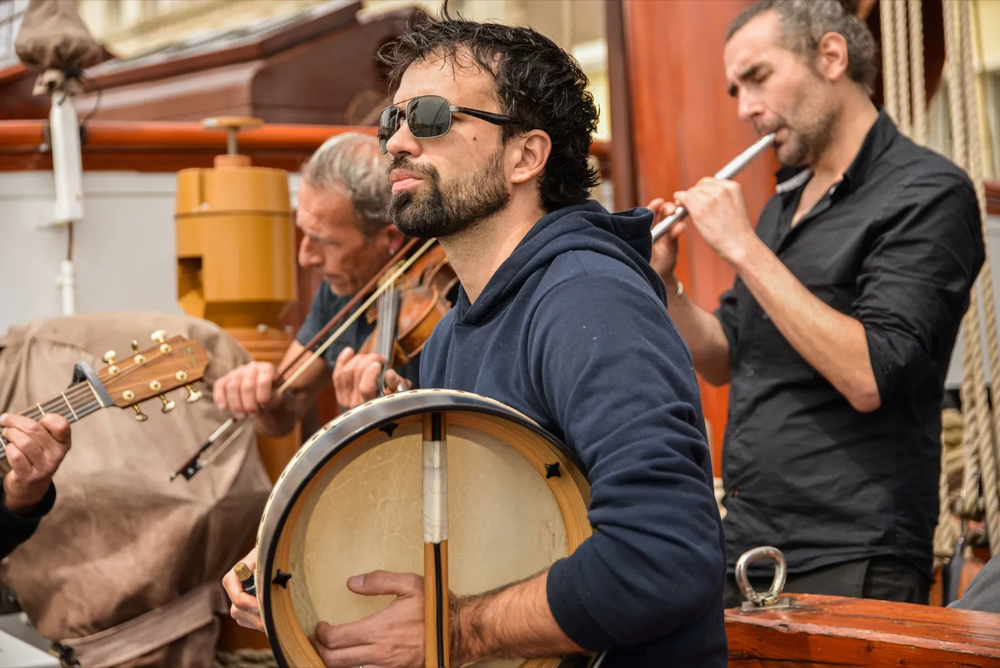 Trois musiciens interprètent un morceau à bord d’un bateau : l’un joue du violon, l’autre du tambourin, et un troisième de la flûte.