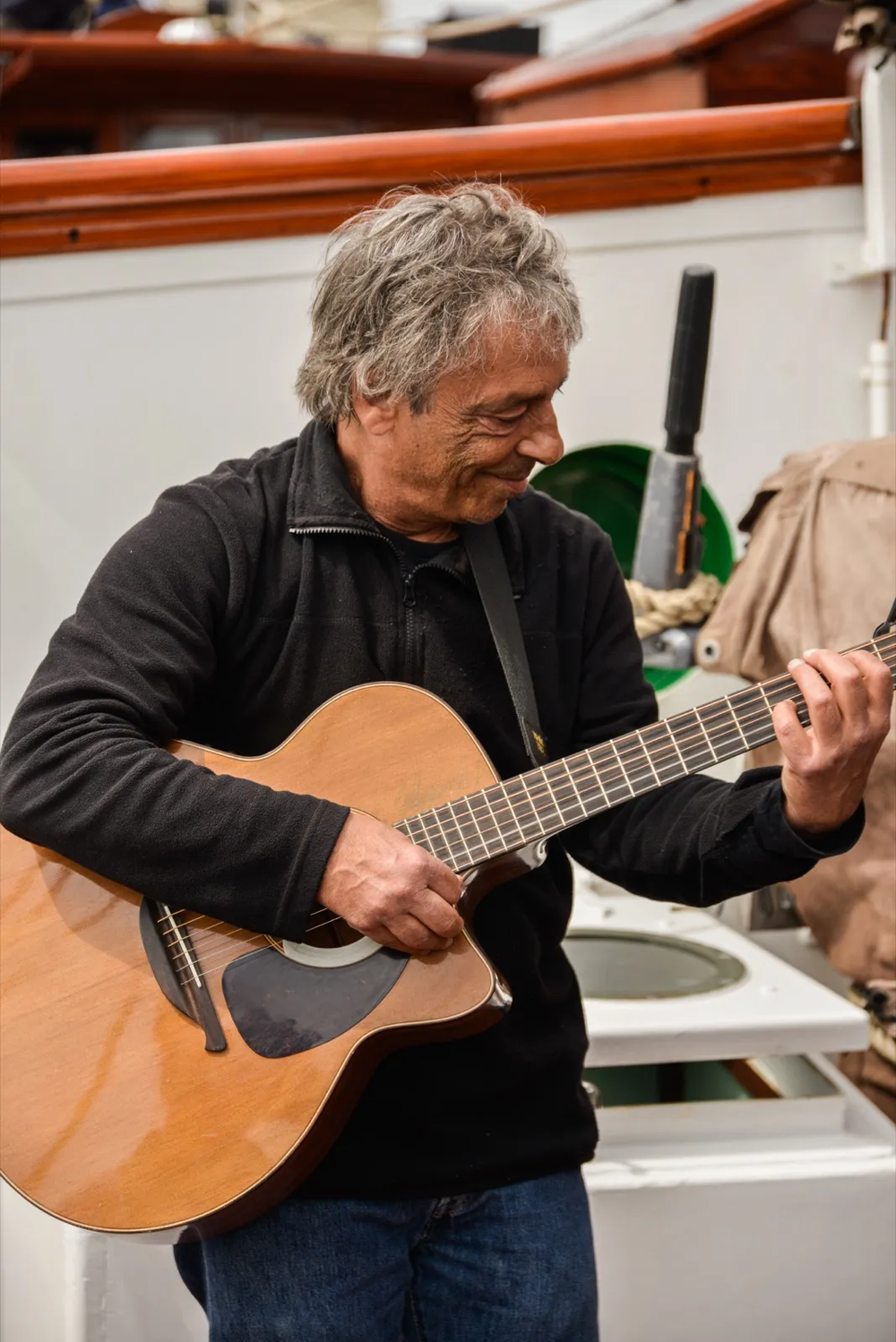 Un musicien aux cheveux gris joue de la guitare acoustique avec concentration sur le pont d’un bateau en bois.