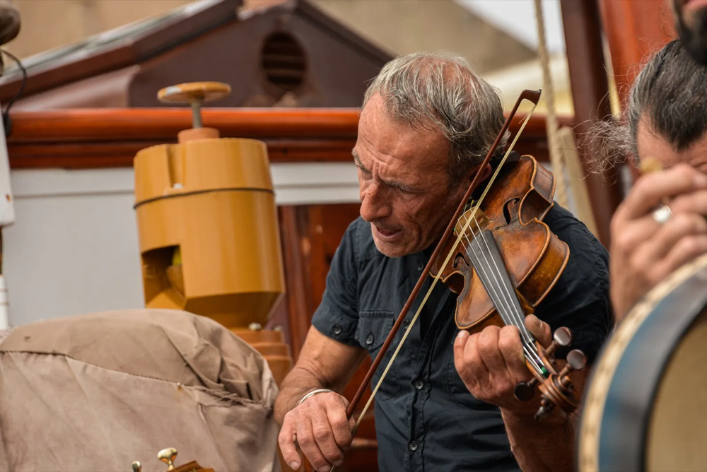 Un homme d’un certain âge joue du violon avec intensité sur le pont d’un voilier, accompagné de musiciens en arrière-plan.
