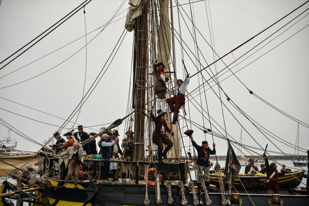 Des marins grimpent dans les cordages d’un grand voilier en bois, vêtus de tenues d’époque, lors d’un événement maritime.