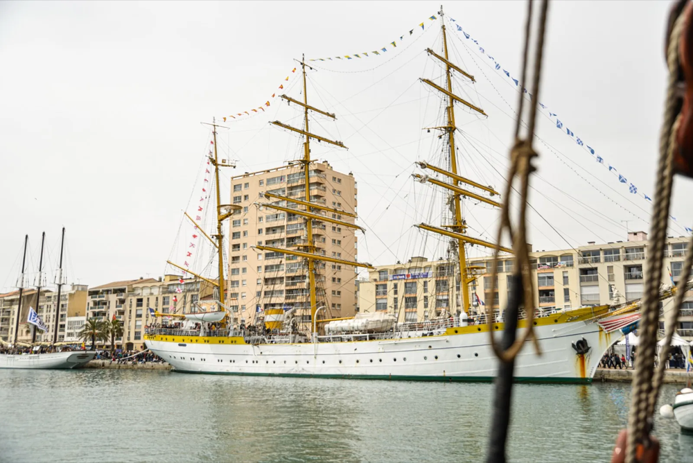 Un grand voilier à quai, orné de fanions colorés, se détache sur un fond de bâtiments modernes en bord de port.