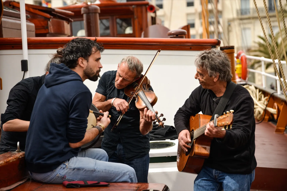 Quatre musiciens assis en cercle sur un pont de bateau, jouant du violon, de la flûte et de la guitare dans une ambiance chaleureuse.