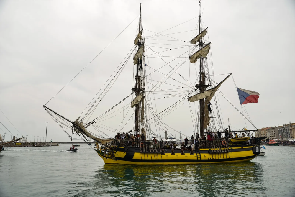 Un grand voilier aux couleurs jaune et noire, pavillon russe hissé, navigue tranquillement sur les eaux calmes du port.