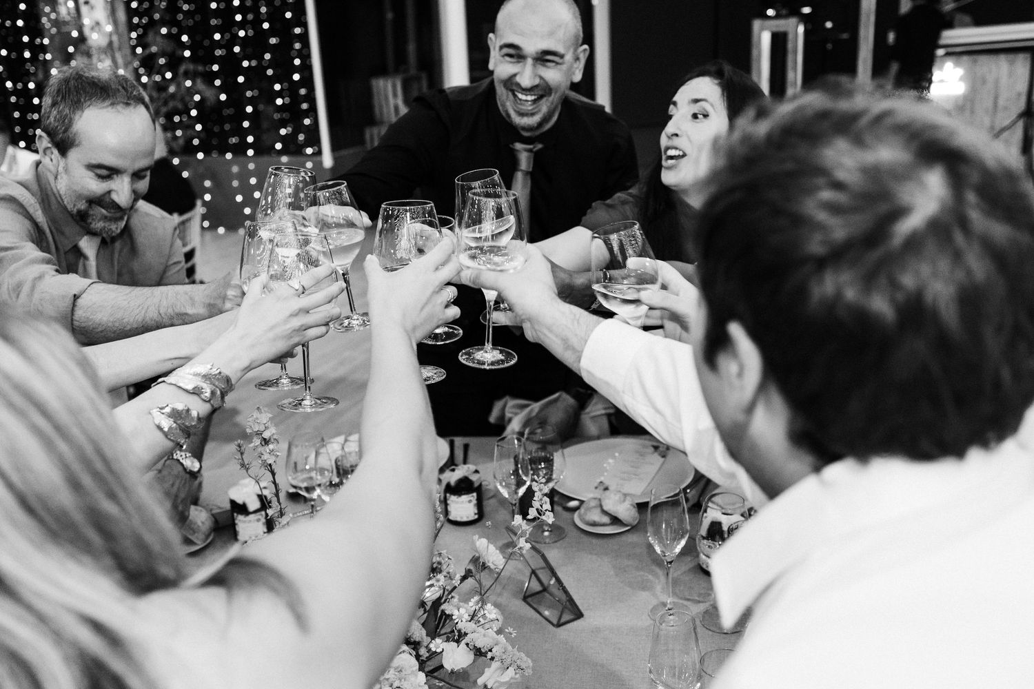 Wedding guests laughing and socializing during speeches at a reception with twinkle light backdrop.