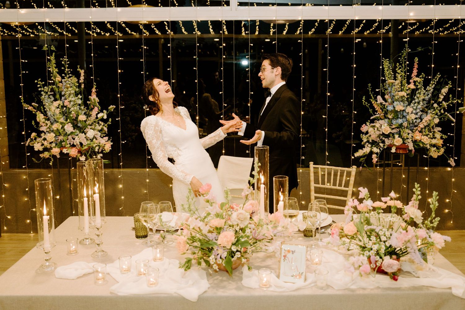 Elegant wedding reception table setting with floral centerpieces and hanging string lights creating a romantic atmosphere.