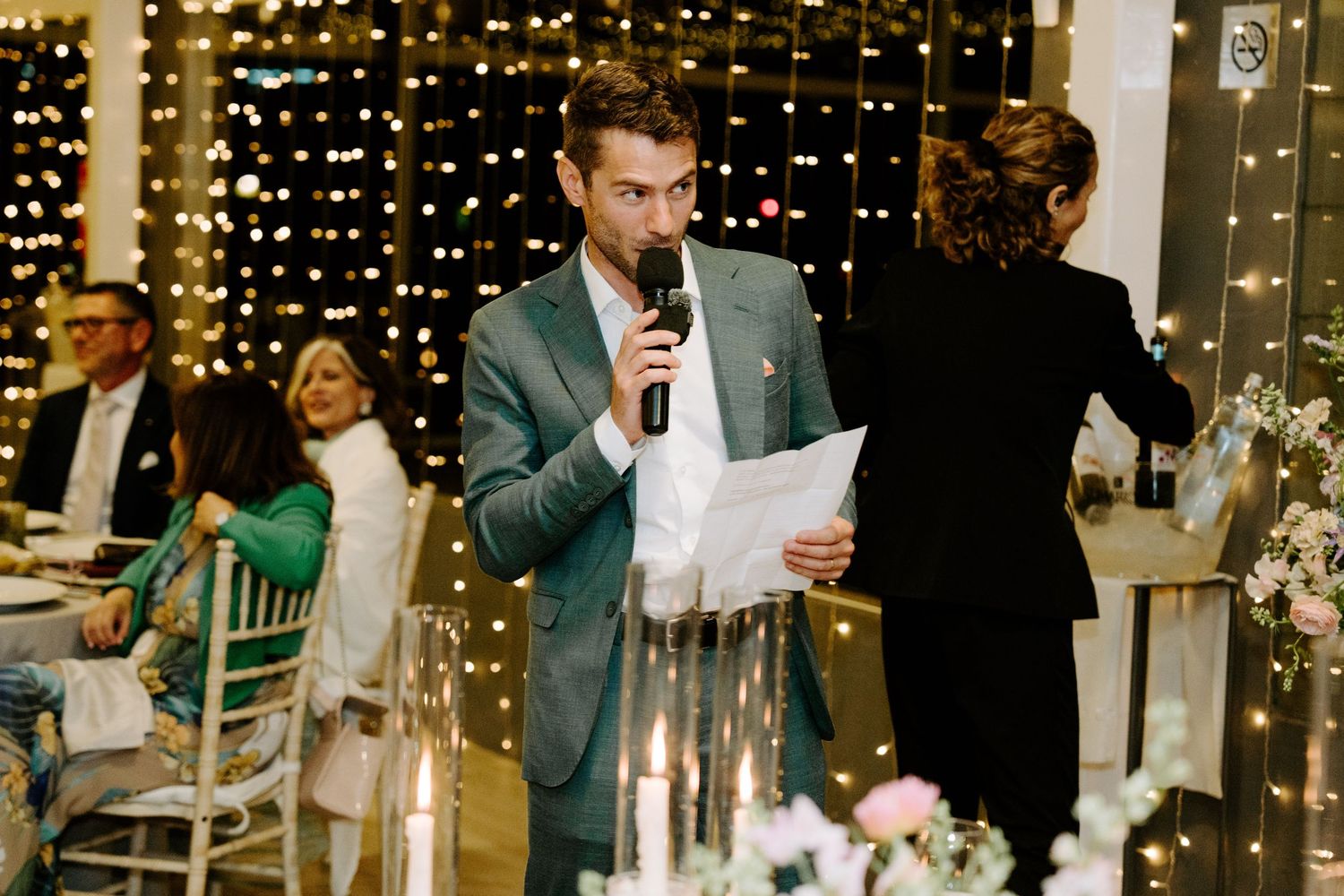Wedding guests listening to speeches during an indoor reception with string light backdrop.