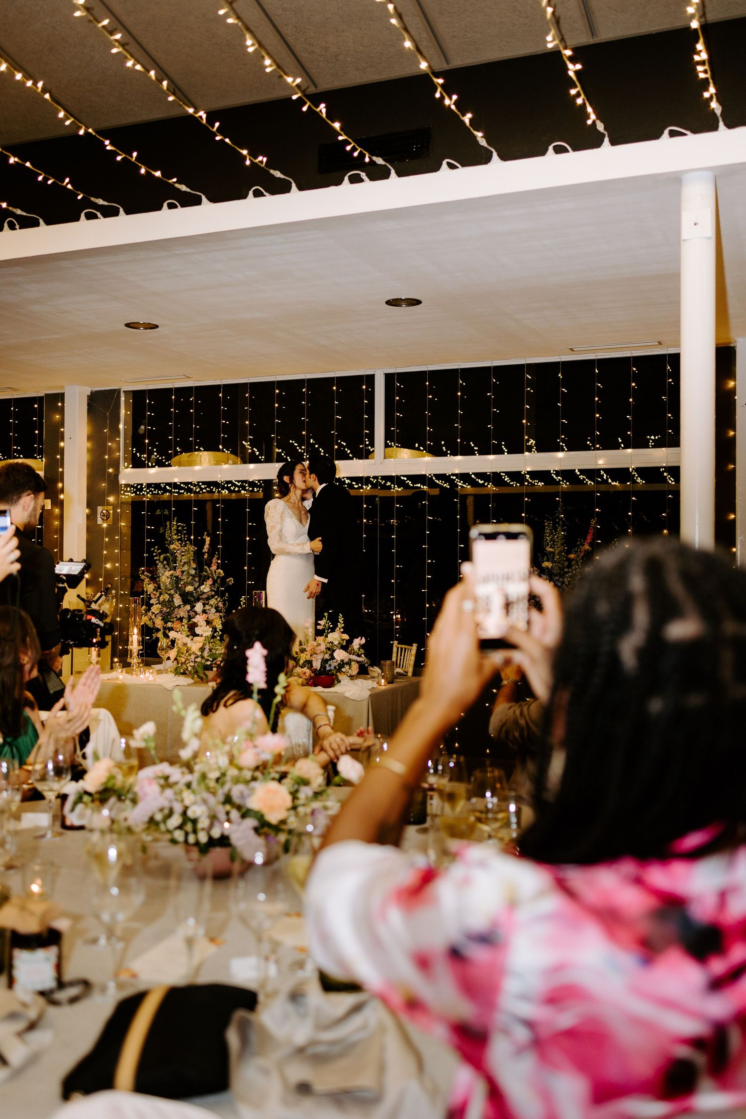 Wedding reception hall with string lights and decorated tables viewed from a distance.