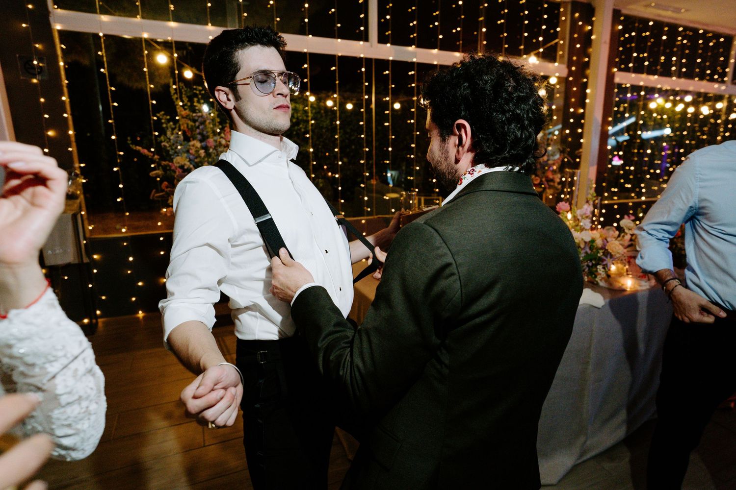 Wedding guests dancing and celebrating on a dance floor illuminated by string lights and colorful lighting effects.