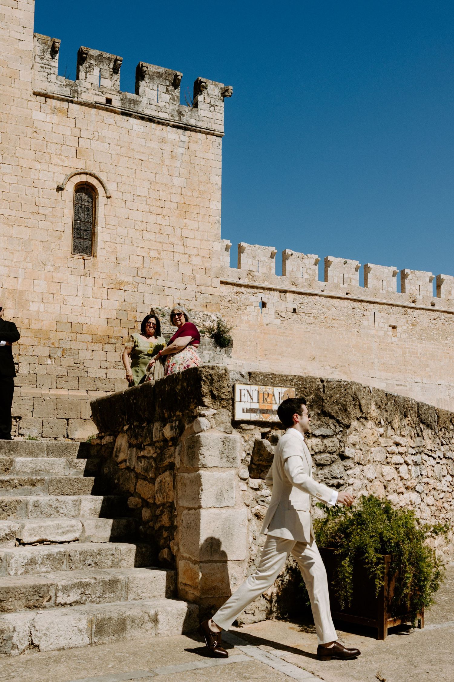 Guests gather outside a historic stone castle with crenellated walls and archways under bright blue sky.