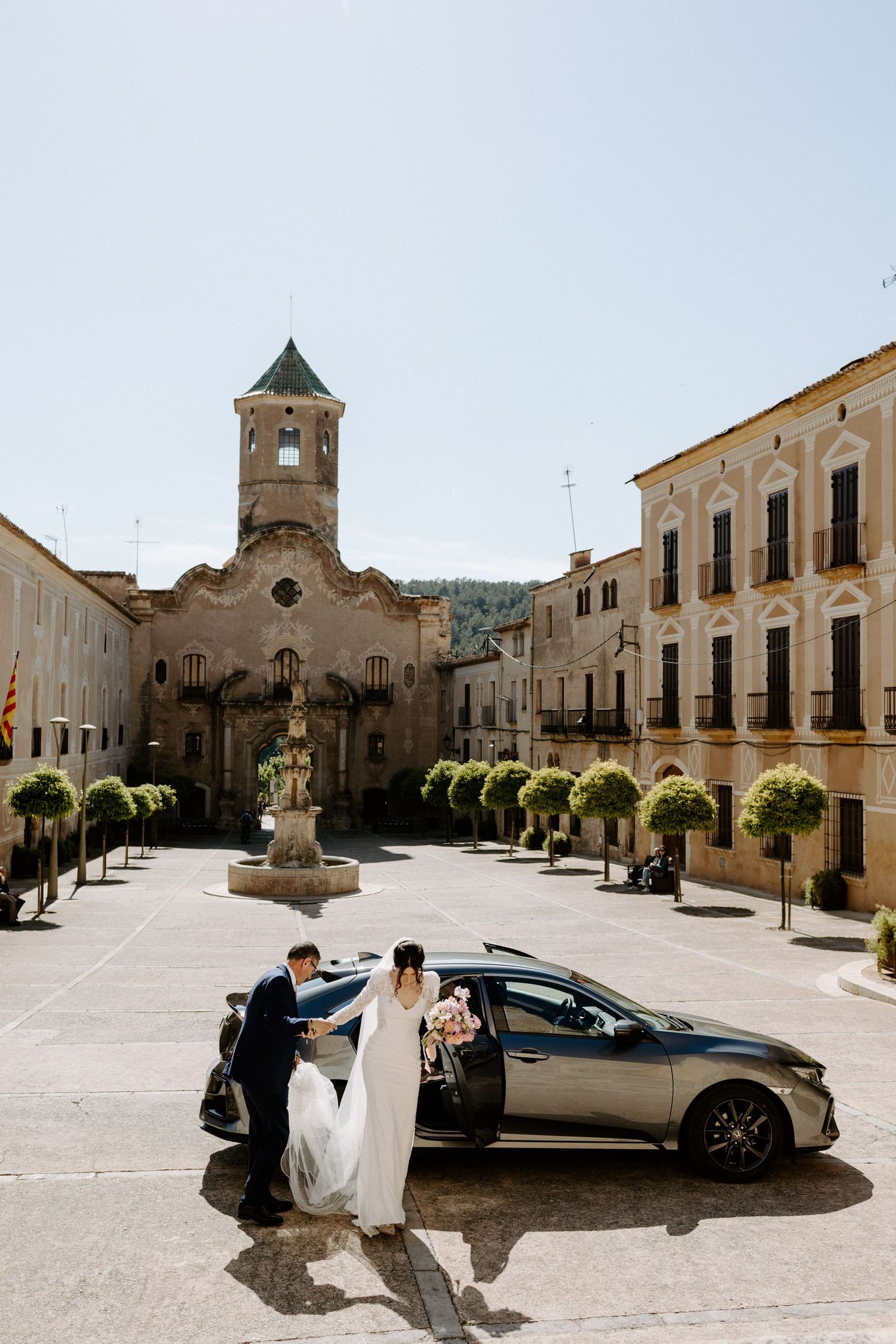 Wedding party and luxury car in front of historic Mediterranean town square and church.